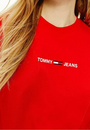 Punainen puuvillapaita, jossa lukee "TOMMY" valkoisella ja "JEANS" valkoisella, ja niiden välissä on sininen ja punainen emblem. Sujuva pinta, pyöreä pääntie.