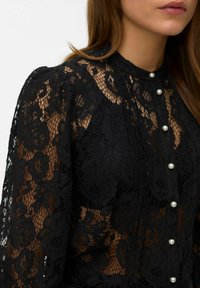 Sort blondebluse med blomsterprint, høj hals og perleknapper ned foran. Den gennemsigtige tekstur tilføjer dybde til designet.