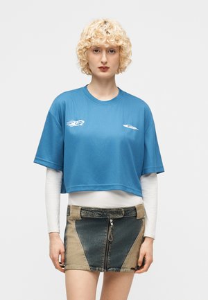 Ung person med krøllet blondt hår iført en blå crop-top T-shirt over en hvid, langærmet top og en tofarvet denim mininederdel med lynlås foran.