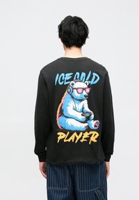 Zwart shirt met lange mouwen met een kleurrijke afbeelding van een ijsbeer die een koptelefoon draagt en een gamecontroller vasthoudt, met de tekst "ICE COLD PLAYER."