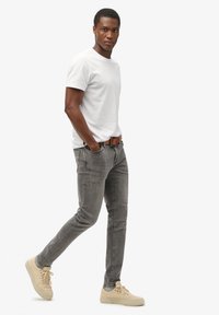 Jeans de denim gris con un corte ajustado, combinados con una camiseta blanca de cuello redondo. El modelo lleva zapatillas beige y un cinturón de cuero marrón.
