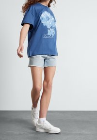 Niebieska bawełniana koszulka z kwiatowym nadrukiem i napisem „Levi's”, zestawiona z jasnymi krótkimi spodniami jeansowymi i białymi trampkami. Casualowy krój i design.