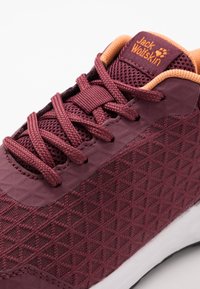 Close-up of maroon Jack Wolfskin sneaker with textured fabric, matching laces, and orange cushioned collar on a white sole.