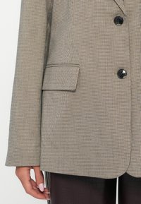 Marynarka z materiału teksturowego w brązowo-beżowy wzór w kraty. Posiada klapę typu notch, dwa guziki i jedną kieszeń na piersi.