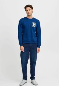 Plavi pamukasti dukserica s okruglim izrezom, rebrastim manžetama i velikom vezenom oznakom "B" na lijevoj strani prsa. Uparen s tamnim trapericama.