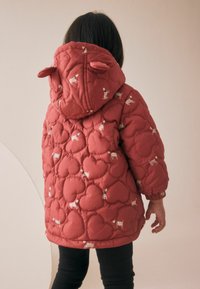 Veste matelassée rouge avec un motif en forme de cœur et de petits motifs de chiens. Elle possède une capuche avec des oreilles et des poignets élastiques. Portée sur des leggings noirs.