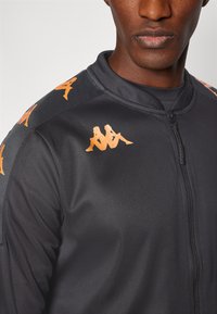 Veste de sport noire avec zip, dotée d'accents de logo orange sur les épaules et la poitrine. Tissu lisse avec un design ajusté.