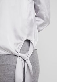 Satin top in lichtgrijs met een zijstrikdetail, voorzien van een ronde uitsparing aan de onderkant en een gladde textuur, gecombineerd met een grijze gebreide ondergrond.