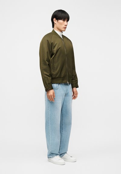 Νεαρό άτομο που στέκεται φορώντας μια φοδραρισμένη μπουφάν bomber σε ελιά πράσινο χρώμα με φερμουάρ, ανοιχτό μπλε τζιν με φαρδιά μπατζάκια, λευκά αθλητικά παπούτσια και λευκό ζιβάγκο.