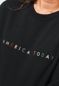 Musta huppari, jossa on monivärinen kirjailtu teksti "AMERICA TODAY" leikkisässä, pyöristetyn muotoisessa fontissa. Pehmeä puuvillamateriaali, hieman rento fit.