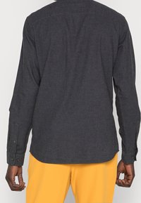 Muž oblečený v tmavě šedé košili s dlouhými rukávy a hořčicově žlutých kalhotách, zobrazený zezadu s uvolněnýma rukama podél těla.