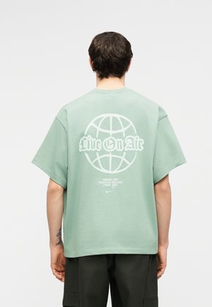 Hombre con cabello corto y oscuro que lleva una camiseta oversize verde claro con un globo terráqueo blanco y el texto "Live On Air" en la parte posterior.