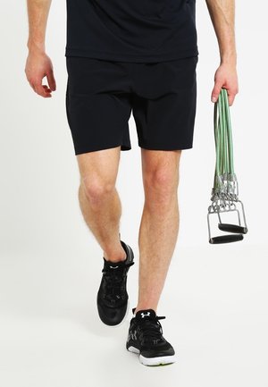 Mand iført sorte shorts og sneakers holder grønne træningselastikker med sorte håndtag, går mod en hvid baggrund.