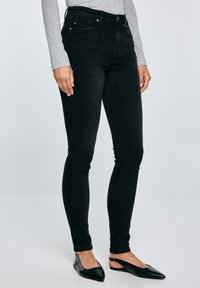 Persona valkā pieguļošas melnas džinsu bikses, pelēku garroku augšu un melnas, ar piespiedumu uz priekšu vērstām kurpēm ar siksniņu aizmugurē, stāvot pret pelēku fonu.