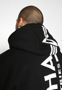 Felpa nera con cappuccio e una grande grafica bianca sul retro, con testo e strisce. Il materiale sembra morbido con una finitura testurizzata.