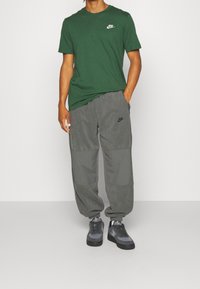 Osoba oblečená v zeleném tričku Nike, šedých teplácích Nike a šedých teniskách Nike stojící na jednobarevném pozadí.