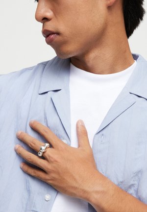Homme en chemise bleu clair et tee-shirt blanc, main sur la poitrine montrant une bague argentée brillante avec trois sphères connectées au doigt.