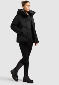 Žena u crnoj quilted puffer jakni, crnim tajicama i crnim čizmama do koljena, stoji s jednom rukom u džepu jakne.