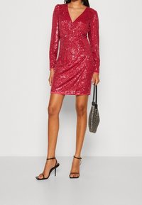 Röd paljettklänning med långa ärmar, V-ringning, kroppsnära siluett och minilängd. Matchad med svarta klackar med remmar och en liten silverväska.