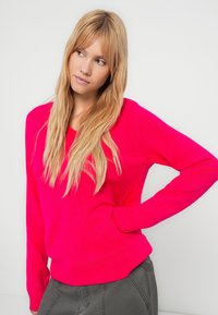 Gestrickter V-Ausschnitt-Pullover in leuchtendem Pink, mit gerippten Bündchen und Saum, mit glatter Textur. Getragen zu grauer Hose. Hintergrund ist einfarbig.