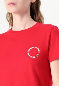 Rød bomull T-skjorte med en rund hvit logo som har teksten "TOMMY JEANS NEW YORK CITY" og hjerteaksenter på venstre brystområde. Korte ermer.