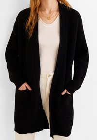 Cardigan en tricot noir avec devant ouvert, manches longues et deux poches avant. Tissu texturé et coupe décontractée. Porté sur un haut de couleur claire.