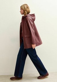 Bordeaux regnjacka med huva, tryckknappar och framfickor, som matchas med lösa mörka jeans och bruna skor.