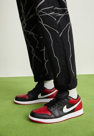 Osoba nosząca czarno-czerwone buty Nike z białym znakiem, białe skarpetki i czarne spodnie z białymi abstrakcyjnymi liniami na zielonej podłodze.