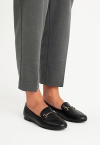 Pantaloni negri din piele cu oアクセoriu decorativ din lanț auriu, asortați cu pantaloni gri, tailored, cu croială dreaptă și lungime până la gleznă.