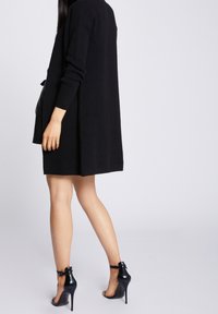 Czarny dzianinowy sweter zapinany na guziki, do kolan, z otwartym przodem i długimi rękawami. Noszony na czarnej spódnicy, zestawiony z czarnymi szpilkami z paskami wokół kostek.