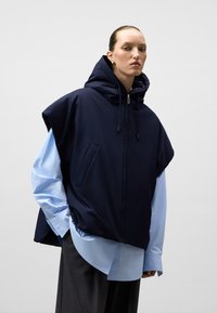 Granatowy bezrękawnik z kapturem, szerokimi wycięciami na ramionach i kieszeniami na piersi, nałożony na jasnoniebieską koszulę. Materiał wydaje się gładki i lekko odbijający światło.