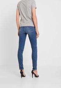 Dünne blaue Jeans mit Kontrastnähten, mittlerem Bund und Gesäßtaschen. Kombiniert mit einem grauen kurzärmeligen Hemd und schwarzen High Heels.