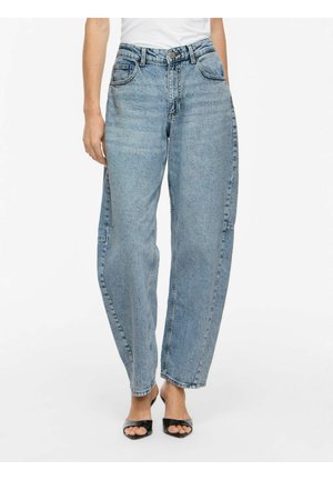 Person iført lyseblå jeans med høj talje og vide ben og sorte åbne hæle, stående mod en hvid baggrund.