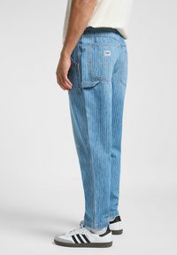 Pantaloni in denim a righe blu chiaro con vestibilità rilassata, dotati di tasche laterali e un'etichetta con logo sul retro. Abbinati a scarpe da ginnastica bianche.
