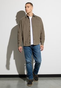 Man walking indoors wearing a brown button-up shirt over white t-shirt, blue jeans, and black boots, casting a shadow on a white wall.