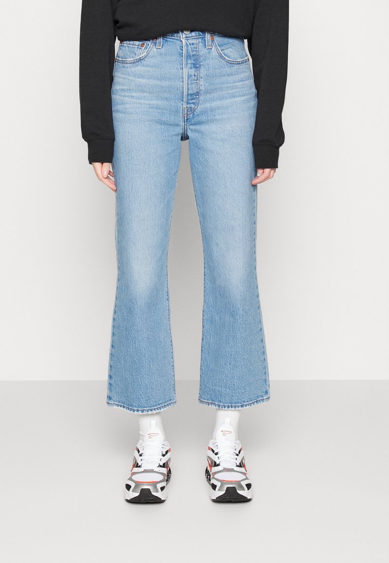 Personne portant un jean flare court bleu clair, un haut noir à manches longues, des chaussettes blanches et des chaussures de sport blanches avec des détails noirs et rouges, debout sur un sol gris.