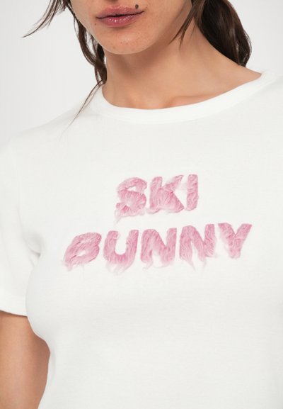 Valge T-särk, millel on tekstureeritud roosad tähed, mis moodustavad sõnad "SKI BUNNY". Kirjatüüp on pehme ja kohev, tõstetud efektiga.