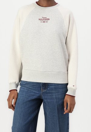 Osoba w jasnoszarej bluzie Tommy Hilfiger z bordowym logo i niebieskich dżinsach, stojąca z ramionami swobodnie opuszczonymi wzdłuż ciała.