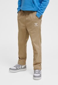 Oseba, oblečena v bež Hummel hlače z rokami v žepih, obuta v bele in sive superge ter modro jopo.