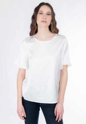 Hvid satinbluse med korte ærmer og udvidede flæsedetaljer, rundet halsudskæring og en glat tekstur. Kombineret med mørke jeans.