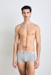 Boxer-briefs en coton gris avec une ceinture blanche arborant le logo "TOMMY HILFIGER" en rouge et bleu. Le modèle se tient devant un fond uni.