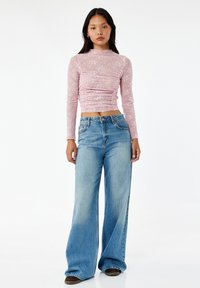 Top de manga larga rosa claro con detalles fruncidos, combinado con jeans de pierna ancha azules. Los jeans tienen un acabado desteñido y un diseño estándar de cinco bolsillos.