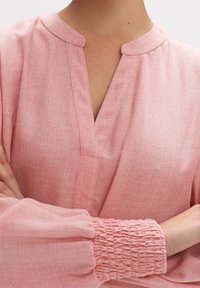 Blouse rose clair avec un col en V, dotée d'un col à bande et de poignets froncés. La texture semble douce et légèrement texturée.