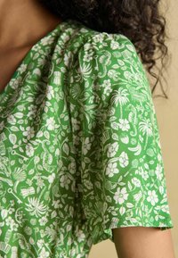 Grünes Stoffoberteil mit kurzen Ärmeln, das ein weißes florales und botanisches Muster mit Seepferdchen, Blumen und Blättern aufweist.