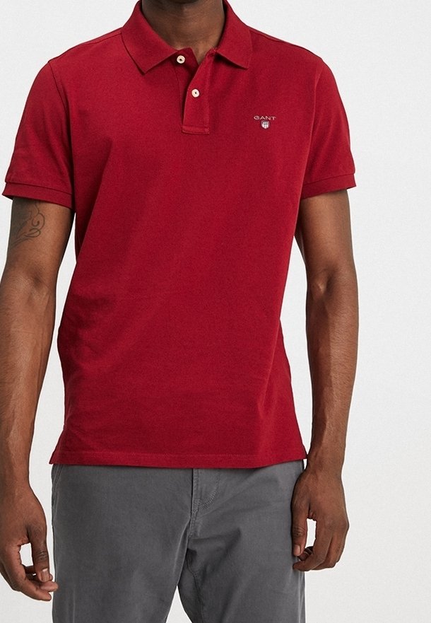 Homme portant un polo rouge uni à manches courtes avec deux boutons et un pantalon gris, debout devant un fond blanc.