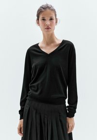 Maglione nero con scollo a V in morbido tessuto a maglia, caratterizzato da maniche lunghe e una vestibilità ampia, abbinato a una gonna nera plissettata.
