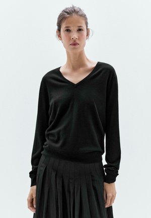 Pull noir en V en tissu tricot doux, avec des manches longues et une coupe décontractée, associé à une jupe noire plissée.