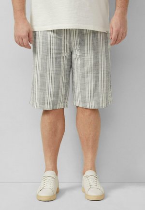 Mann trägt knielange graue und weiße vertikal gestreifte Shorts, weiße Sneaker und ein cremefarbenes Hemd und steht vor einem einfarbigen Hintergrund.