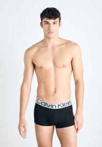 Crni pamučni bokserice s širokim sivoj gumici na kojoj stoji "Calvin Klein" u crnom slovu. Model je bez majice i stoji uspravno.
