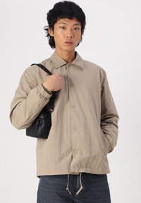Chaqueta de botón color beige claro con puños elásticos y dobladillo con cordón, hecha de una tela suave. Bolsa cruzada negra pequeña sostenida en la mano.